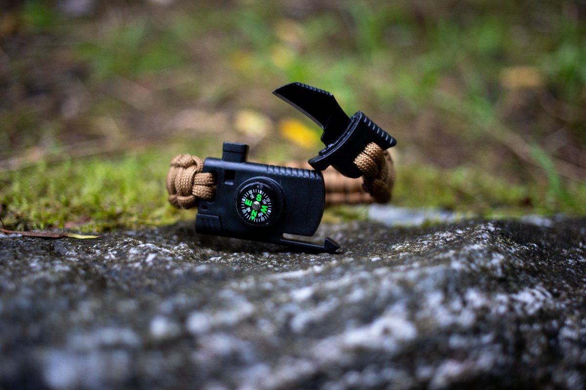 Das Bitehunter 5 in 1 Überlebensarmband / Survival Bracelet hilft in jeder Situation. Es inkludiert einen Feuerstein zum Feuer machen, eine Säge um Äste und Holz zu zersägen, ein Messer um zerschneiden von Seil und Verpackungen, einen Kompass sowie eine Signal Pfeife um Hilfe zu holen. Das Band besteht aus 3 metern paracord und hat gute erfahrungen laut den bewertungen. Hier liegt das braune armband auf einem grauen Stein mit Moos.